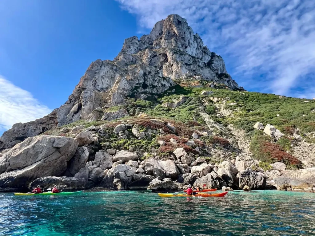 Grupo de kayakistas en travesia de 8 dias remando en kayaks de colores por las aguas turquesas de la costa virgen de Ibiza junto a formaciones rocosas.
